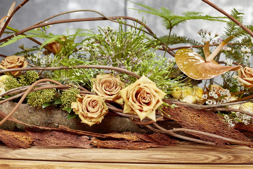 A natural arrangement of beige roses and foliage sitting on top of a wooden log with stems. Made with OASIS® Floral foam.