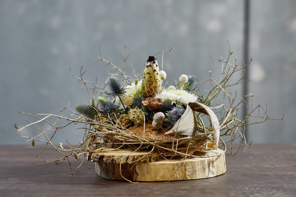 A wood round with a blue, yellow and green arrangement on top. Surrounded with twigs to create a nature inspired look.